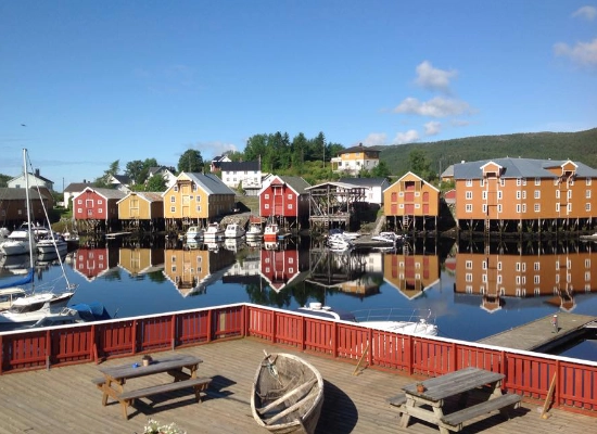 Colorful historic waterfront warehouses on stilts reflected in calm harbor waters at Råkvåg, Trøndelag, Norway, with traditional wooden boat in foreground