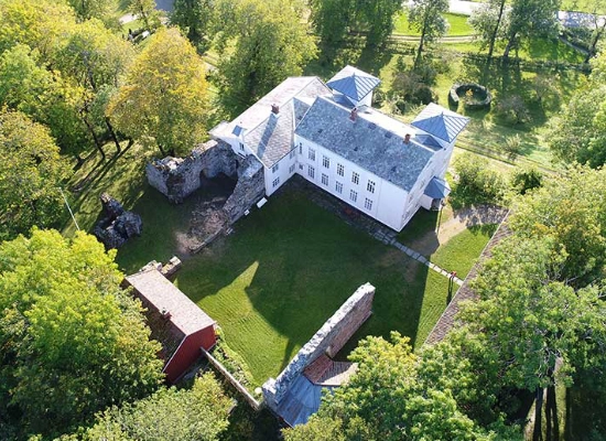 Aerial view of historic white manor house with traditional red outbuildings surrounded by lush green landscape in Rissa, Trøndelag, Norway